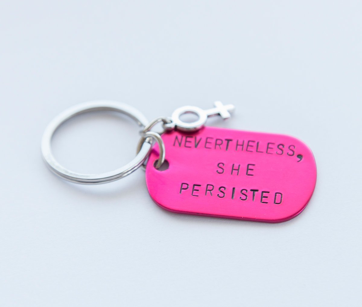 A pink feminist keychain with the phrase 'Nevertheless She Persisted' hand-stamped on a lightweight aluminum tag, attached to a durable stainless steel ring.